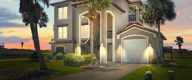 Modern two-story beige house with white trim, two-car garage, and palm trees at sunset